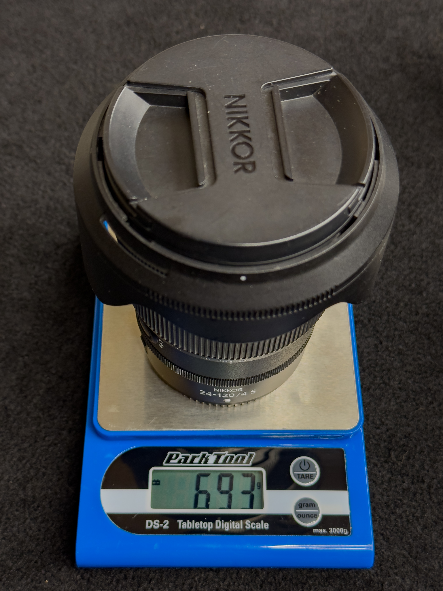 Photo of the Nikon Z 24-120mm f/4 S sitting on a small digital scale displaying its weight, visually underscoring its weight savings compared to traditional f/2.8 professional zooms (including lens hood)